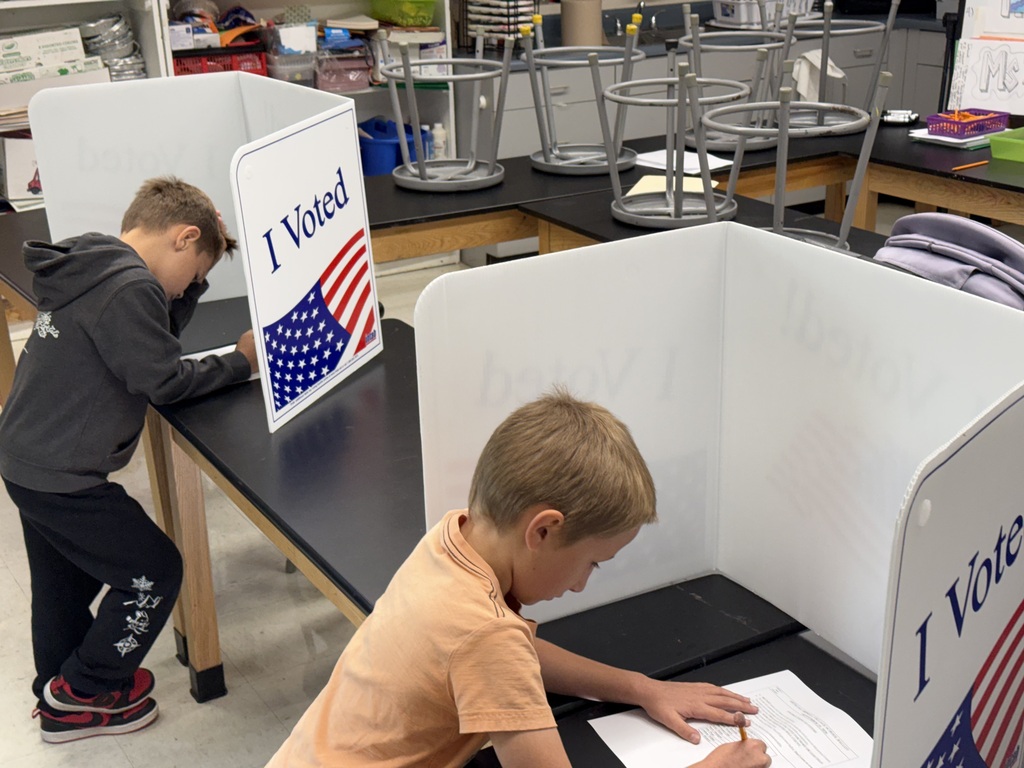 Two students sit at separate classroom desks surrounded by white "I Voted" privacy shields while filling out their paper ballots.