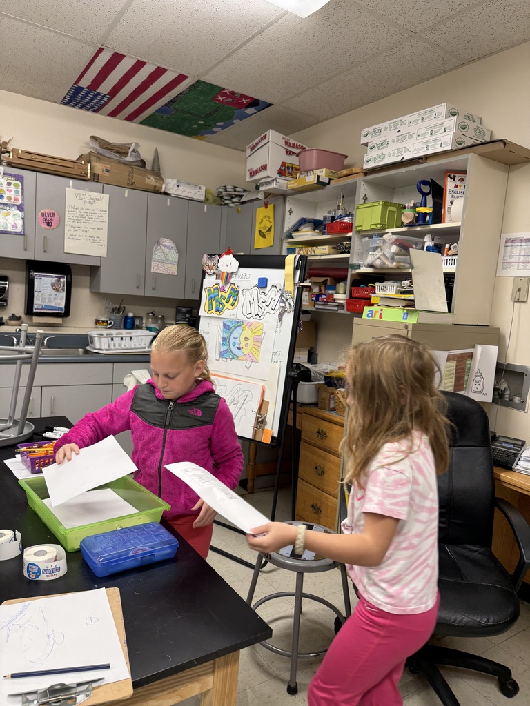 Two young students work at a table in a classroom with art projects, papers, and school supplies surrounding them.