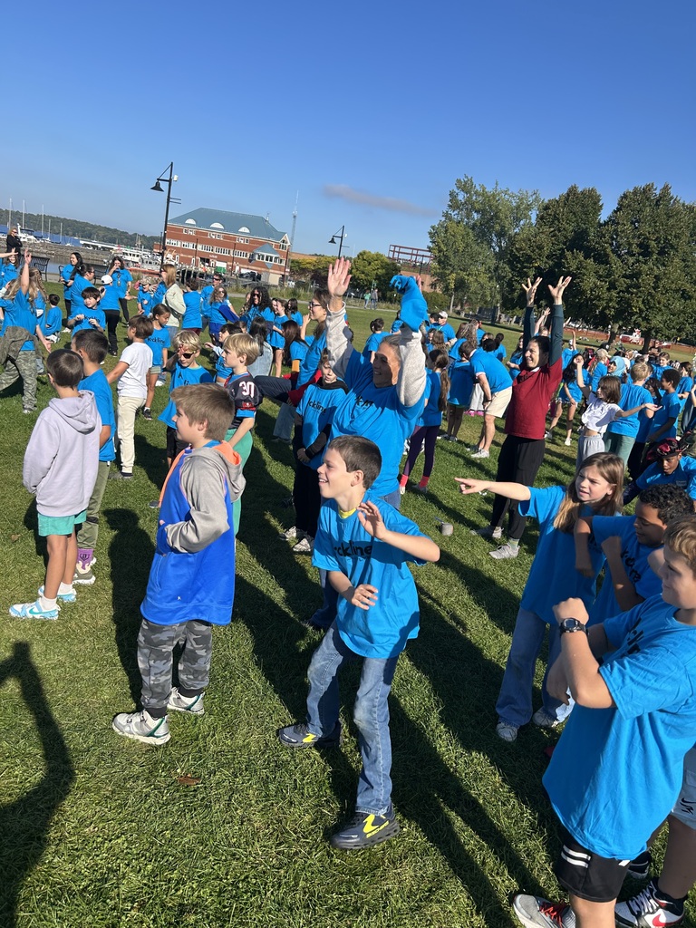 A group of students outdoors on a sunny day wearing matching bright blue t-shirts, lined up on a grassy lawn doing group dance moves and stretching together.