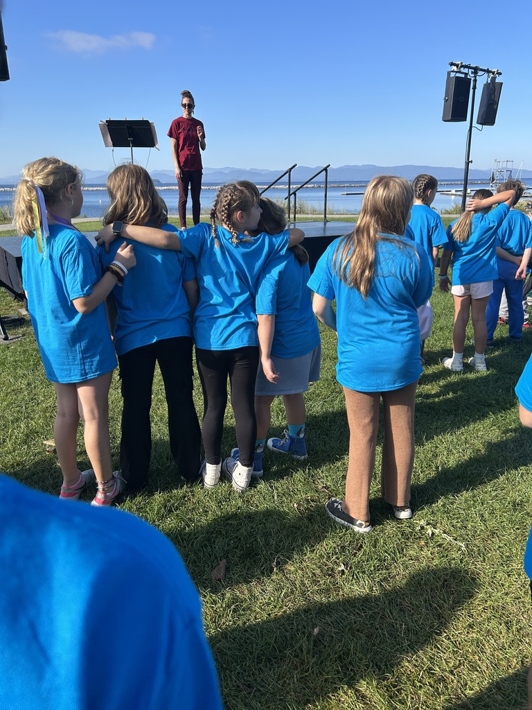 A line of students in bright blue t-shirts standing on a grassy field outdoors next to a body of water, facing toward a speaker standing near a microphone.
