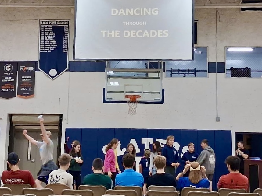 Students gathered on stage during an assembly in the gymnasium while a projection screen above reads "DANCING THROUGH THE DECADES".