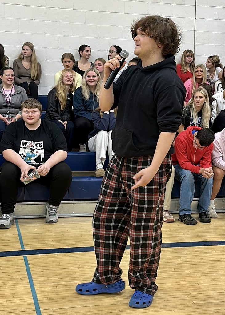 A student in plaid pants speaking into a microphone on the gym floor as fellow students watch from the bleachers.