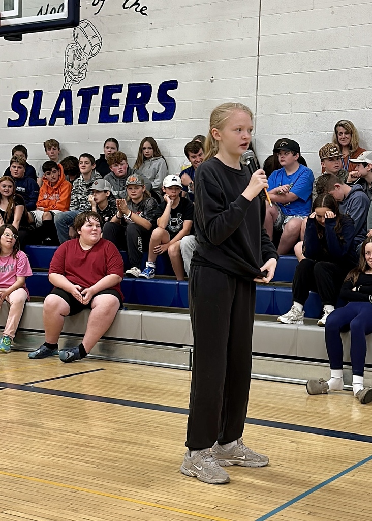 A student standing alone on the basketball court speaking into a microphone with classmates listening in the background.