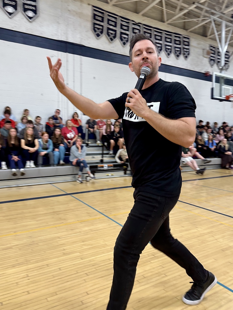 Youth speaker Brandon Lee White holding a microphone and gesturing dynamically while addressing a crowd in the school gym bleachers.