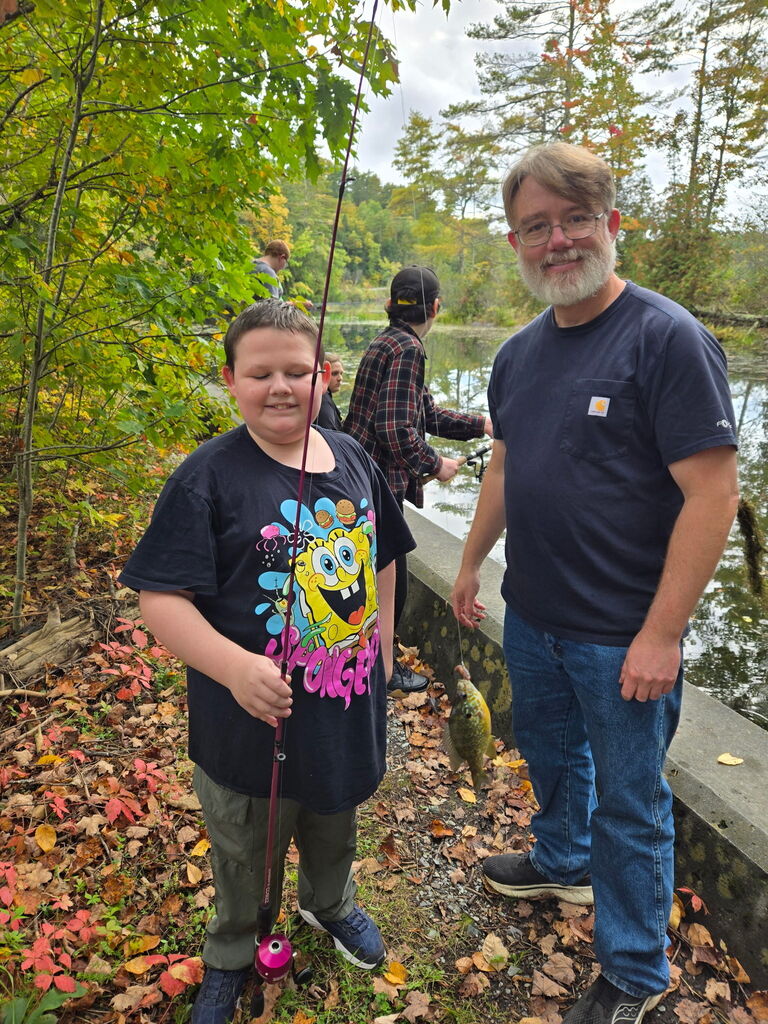 A young boy wearing a SpongeBob t-shirt smiles while holding a fishing pole next to an adult man with glasses and a beard. The man holds up a small sunfish caught on a line along the bank of a wooded pond.