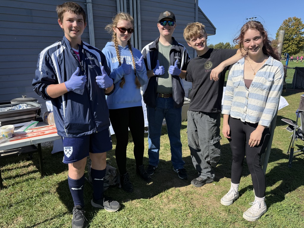 Five students standing outdoors together on a sunny day in front of a catering setup on a lawn. Three of the students are wearing purple disposable gloves and giving thumbs-up signs to the camera.