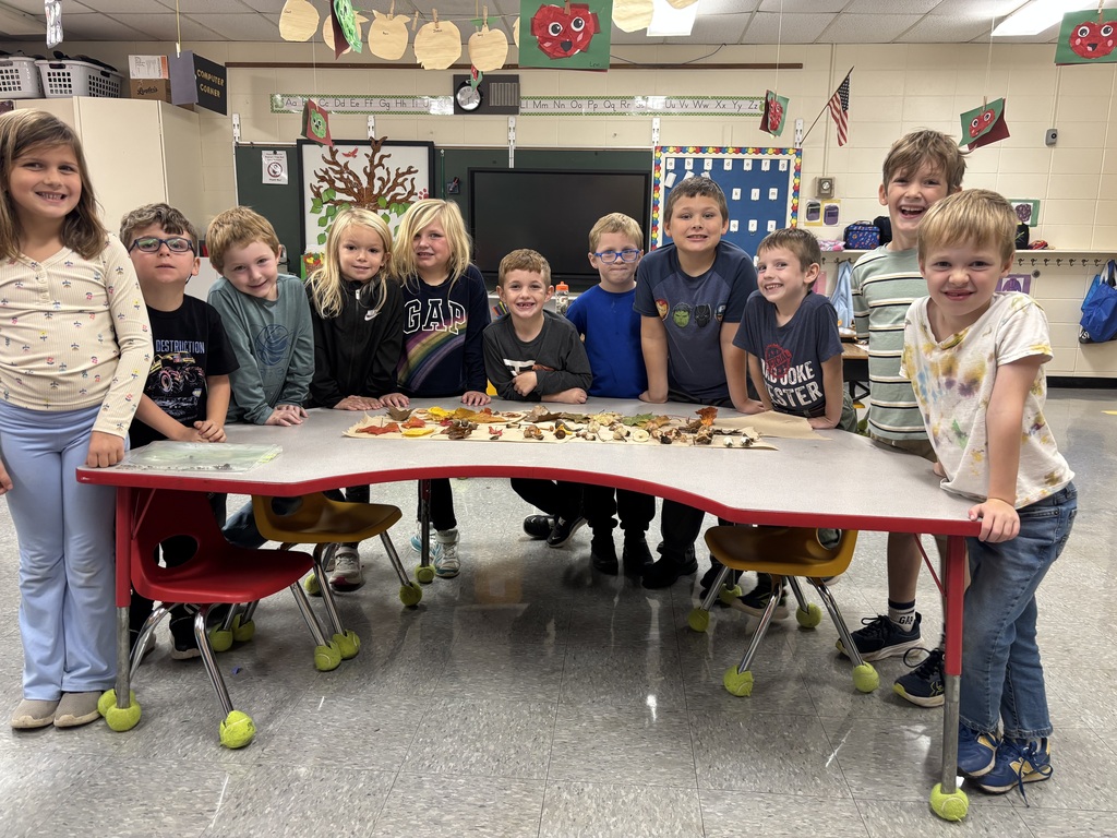 A group of smiling 1st-grade students standing gathered together behind a classroom table that displays their collected fall leaves, acorns, and wild mushrooms.