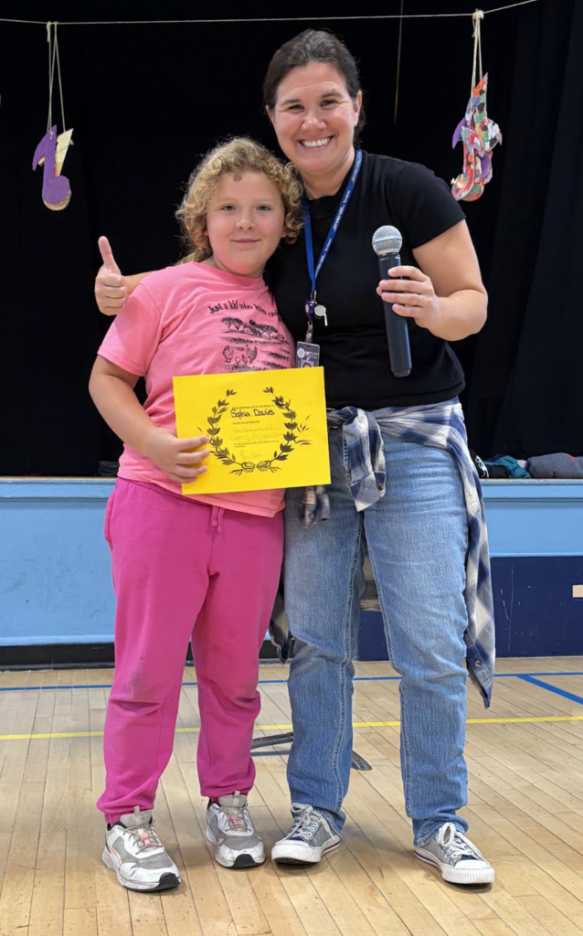 A female staff member holding a microphone standing beside a smiling student giving a thumbs-up while holding a yellow award certificate.