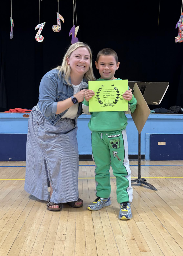 A teacher smiling next to a student holding a bright green award certificate on a school stage.