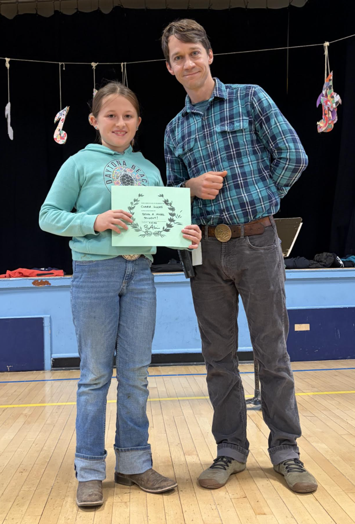 A male teacher standing next to a student holding a light green award certificate on a gymnasium stage.