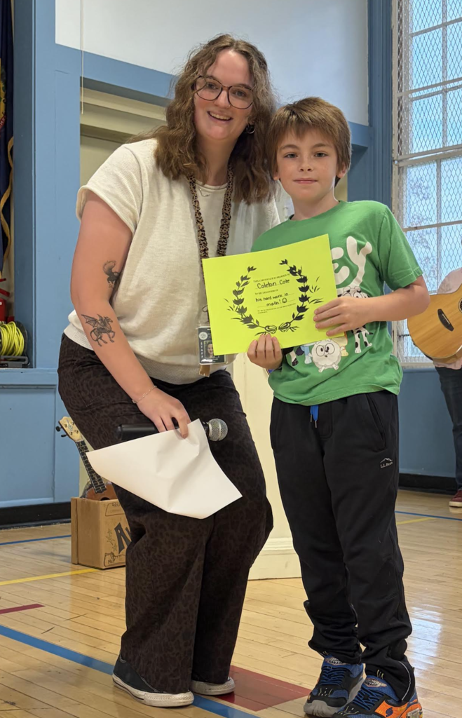 A teacher holding a microphone standing beside a student presenting a bright yellow award certificate.