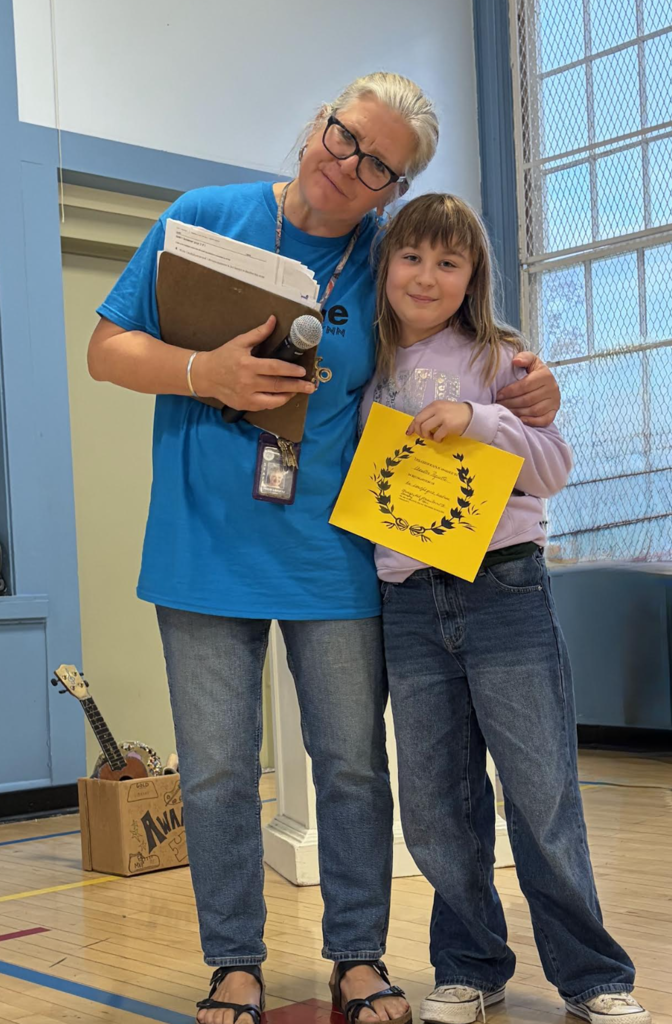 A teacher and student standing together in front of a stage, smiling with a yellow award certificate.