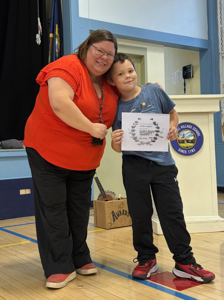 A female staff member posing with her arm around a young boy holding a white award certificate in a gymnasium.