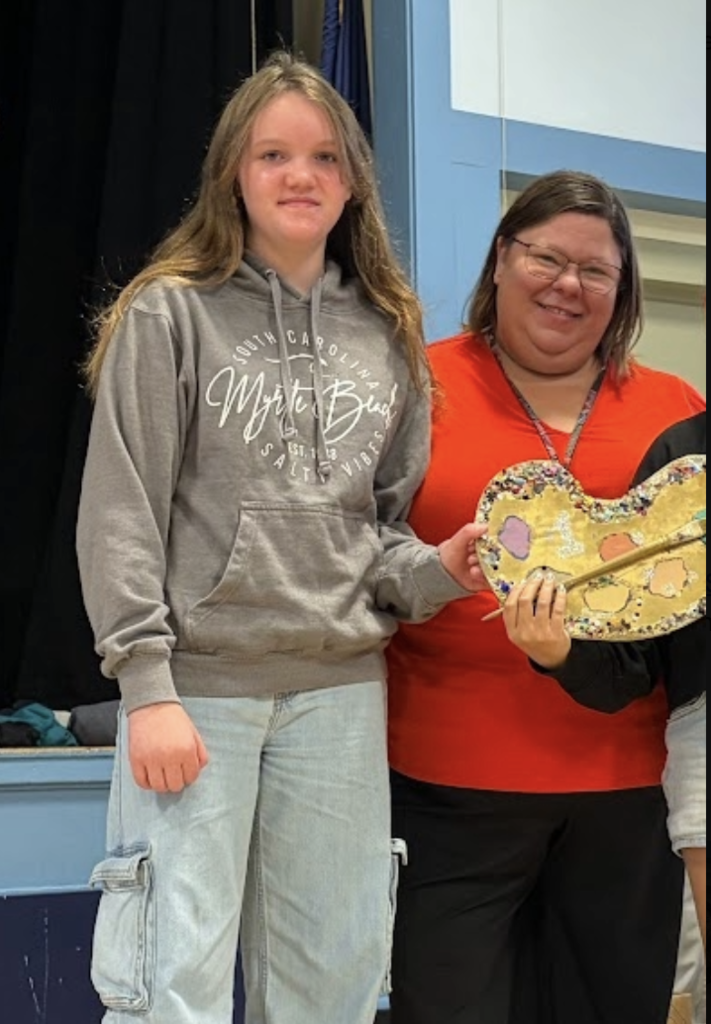 A teacher standing next to a female student who is holding a decorated artist palette trophy.
