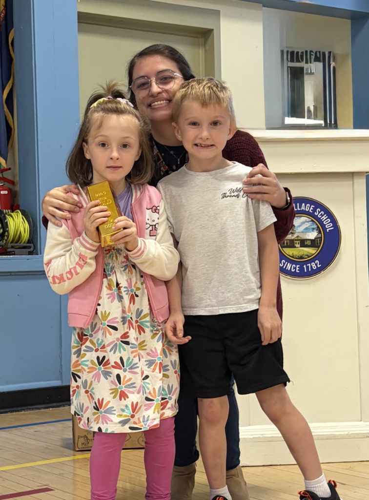 A teacher posing with her arms around two young students, one of whom is holding a gold bar award prop.