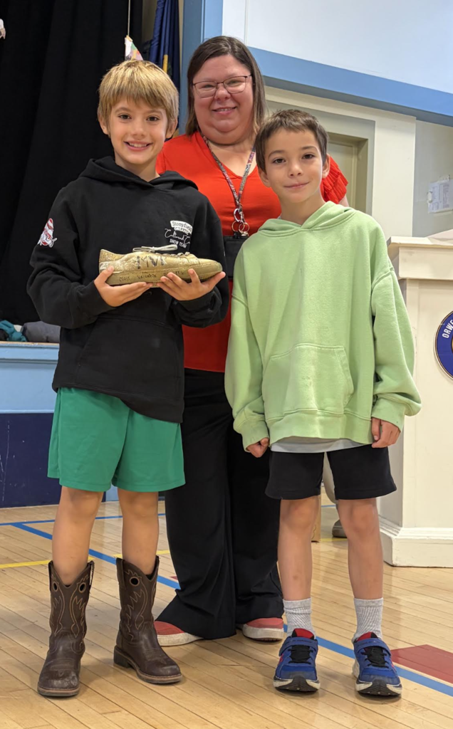 A teacher posing behind two young boys, one of whom is holding up a decorated gold shoe award labeled "MVP".