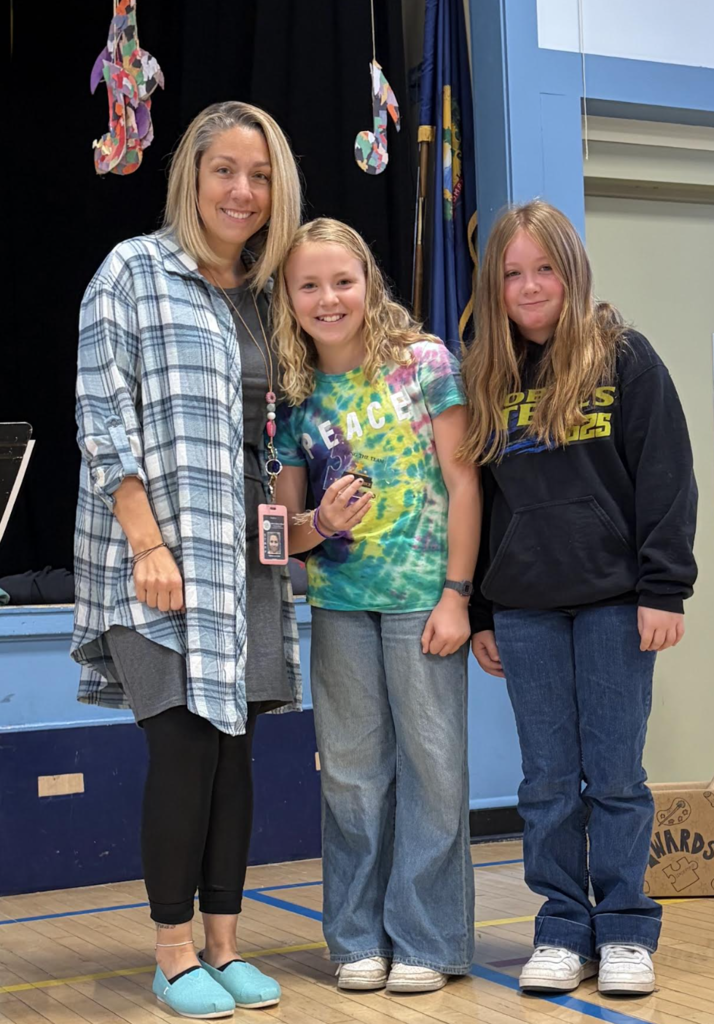 A teacher standing alongside two smiling female students holding a small award on a gymnasium stage.