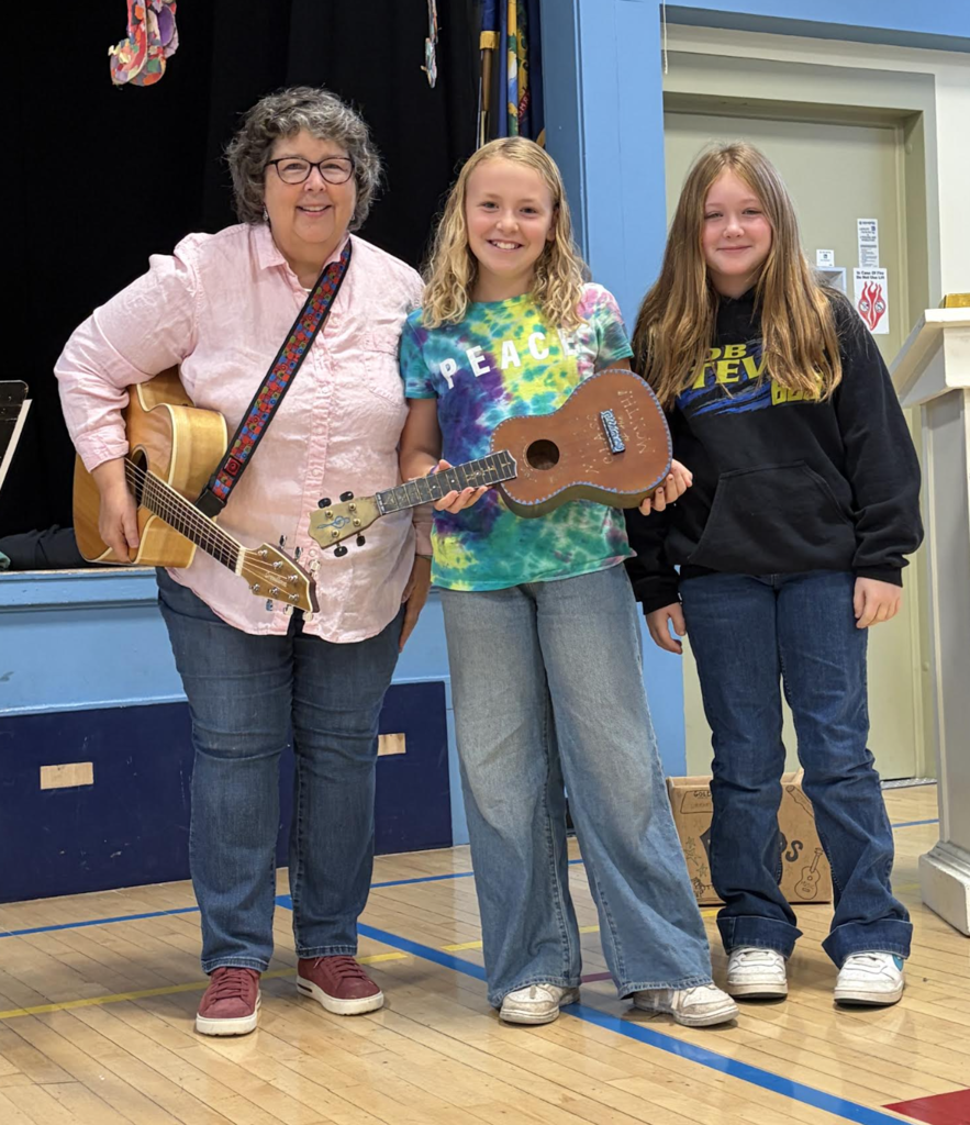 A music teacher holding an acoustic guitar while standing next to two female students displaying a ukulele award.