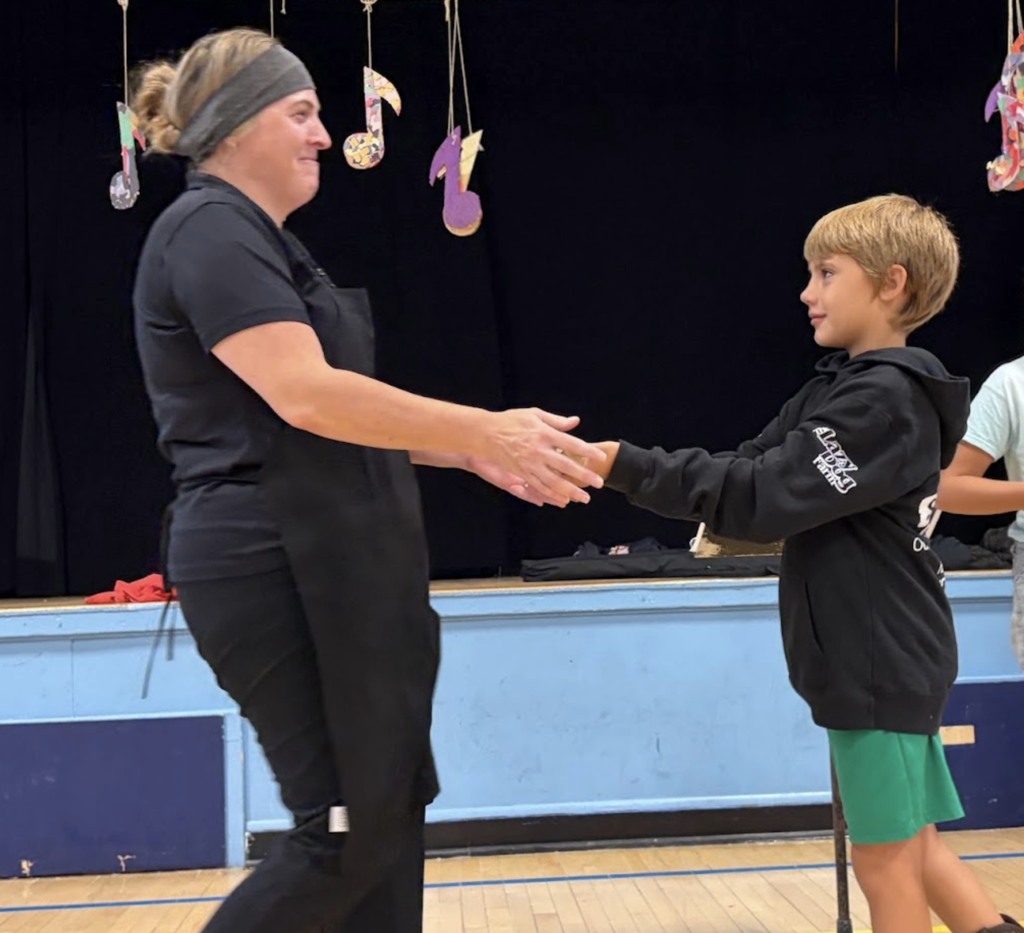 A woman in an apron handshakes a young boy wearing cowboy boots on stage during a school awards presentation.