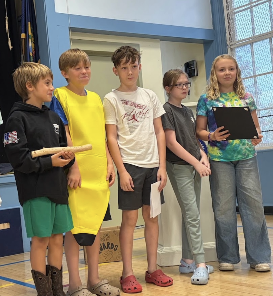 Five elementary students, including one in a banana costume, standing together on a gymnasium stage during a school assembly.