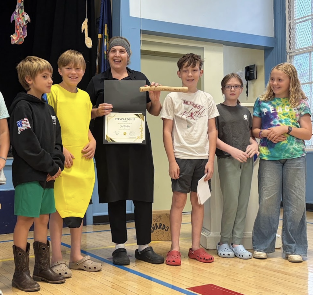 A school chef holding a Stewardship Award certificate stands smiling alongside five elementary students on a gymnasium stage.