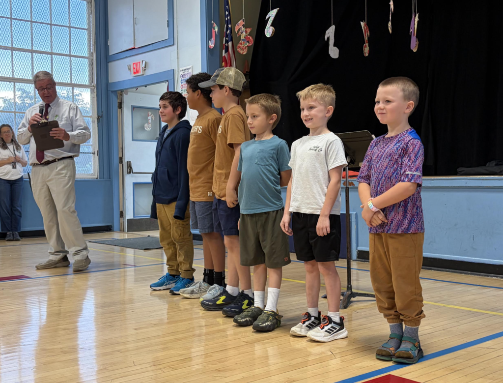 Six young boys standing in a row on a gymnasium floor while a man reads from a clipboard into a microphone during a school assembly.