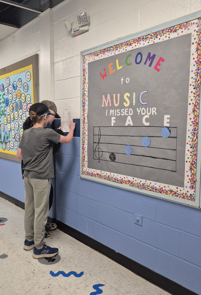 Two elementary students wearing baseball caps look closely at a cast list posted on a blue hallway wall outside a classroom.