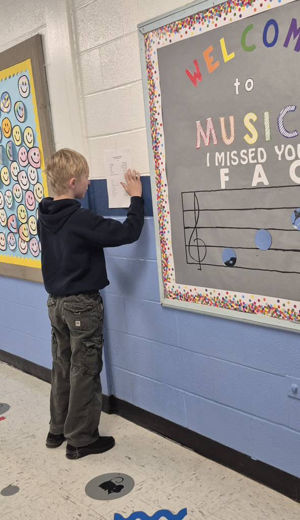 A young student in a dark hoodie and cargo pants reaches up to read the posted cast list on a school hallway wall.