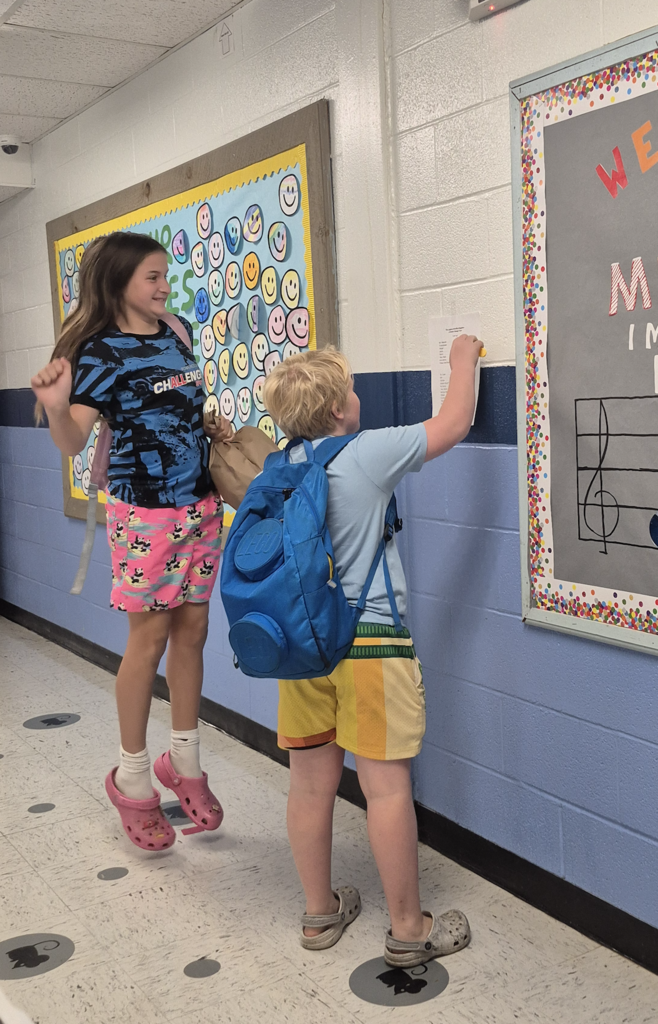 Two elementary students stand together in a school hallway, checking the cast list posted on the wall under a colorful music bulletin board.