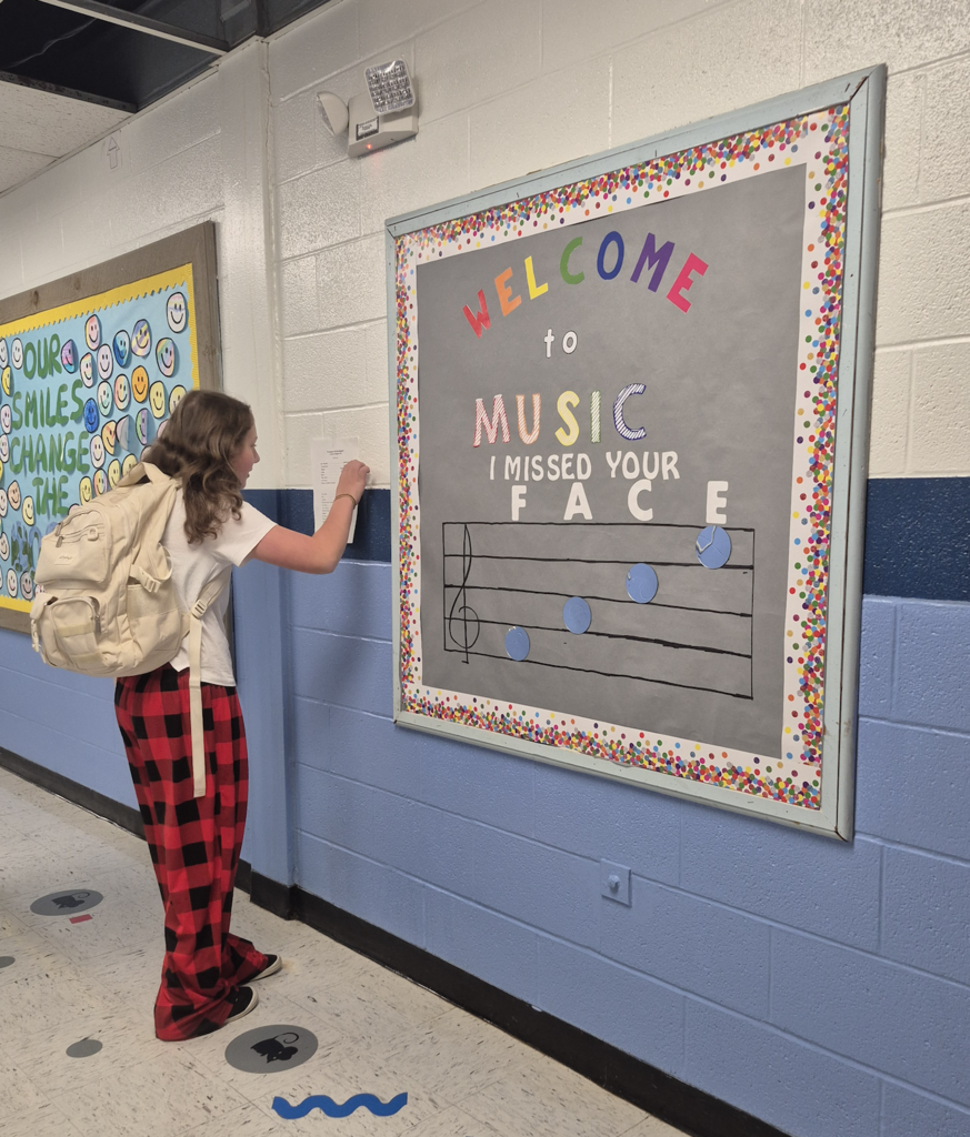 An elementary student wearing red-and-black plaid pants and a backpack stands in a school hallway, looking up at a posted paper on the wall beneath a bulletin board that reads "Welcome to Music".