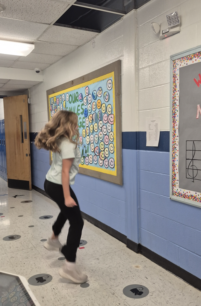 An elementary student with blonde hair walks briskly down a school hallway past bulletin boards and class postings.