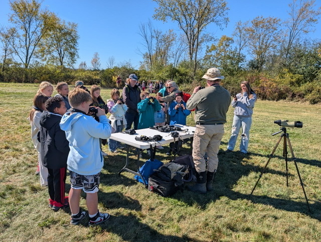 An outdoor field station setup where students use binoculars near a folding table, with a spotter scope on a tripod positioned nearby.