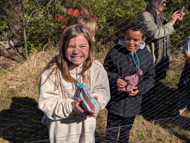Two smiling children outdoors holding small, colorful cloth bird-banding bags close to a mesh netting enclosure.