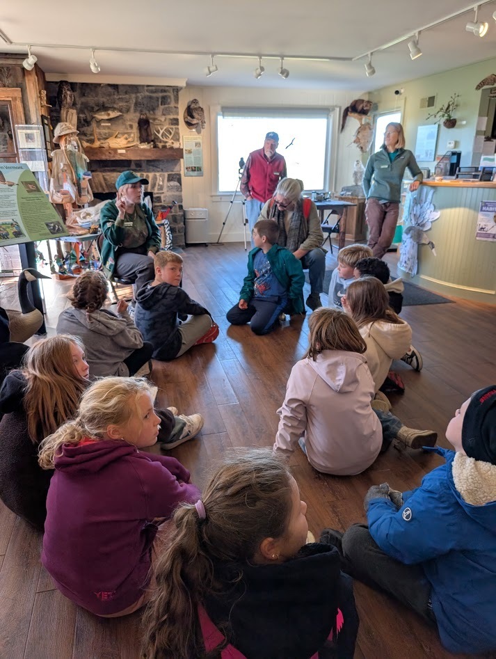 Students sitting on the wooden floor of a nature center room, listening attentively to an instructor giving a presentation near a stone fireplace.