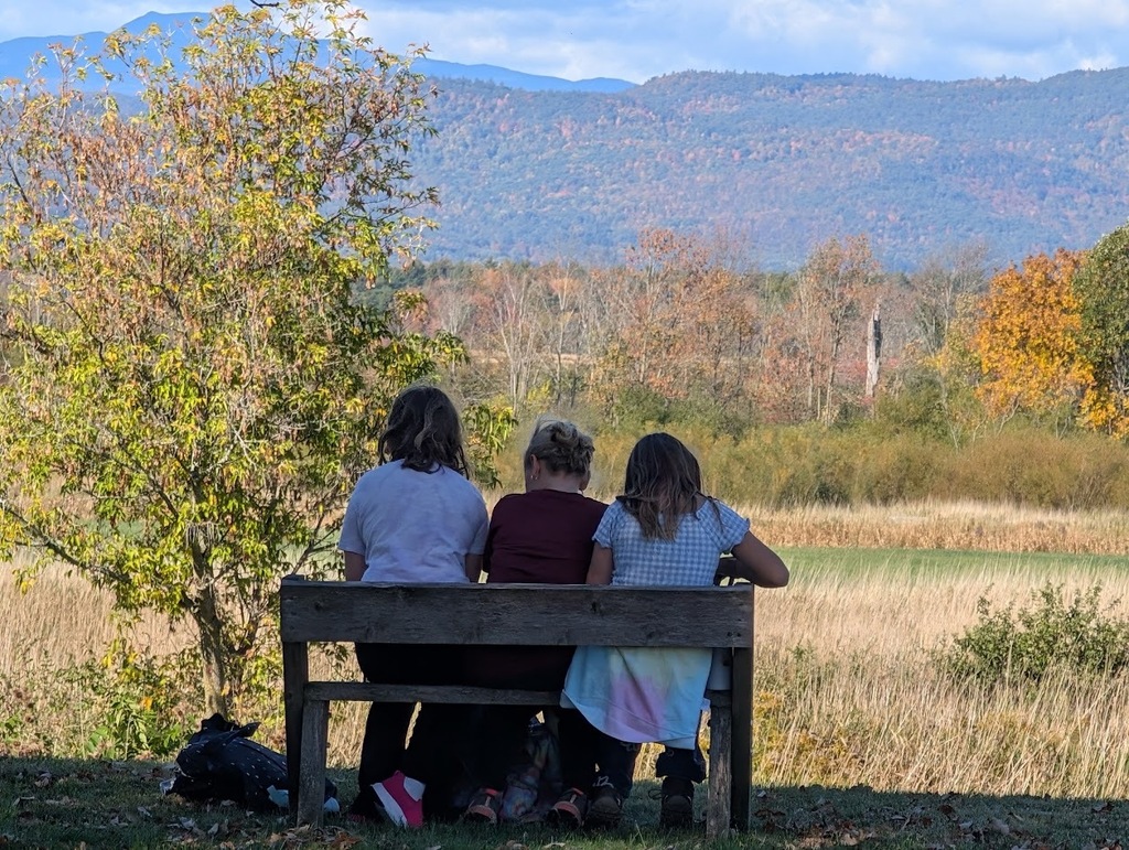 Three students sit side-by-side on a wooden bench, viewed from behind, looking out over an open grassy field toward scenic rolling mountains with autumn foliage.