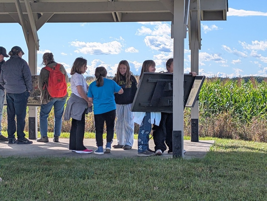 A group of students gathered under an outdoor covered shelter, looking at informational display boards next to a tall cornfield.