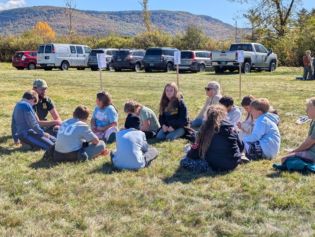 A group of students sitting in a circle on the grass in a sunny field, listening to an instructor, with parked vehicles and hills in the background.