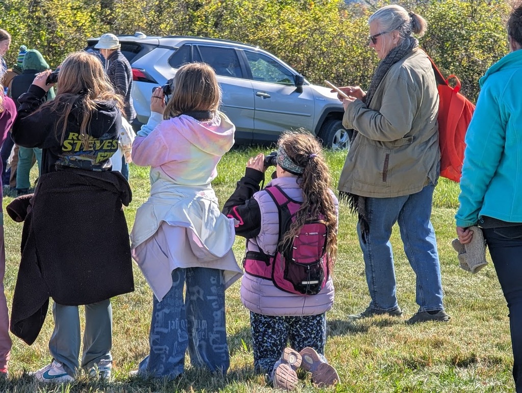 Students standing and kneeling in a grassy field, holding binoculars to their eyes as they observe wildlife alongside an adult supervisor.