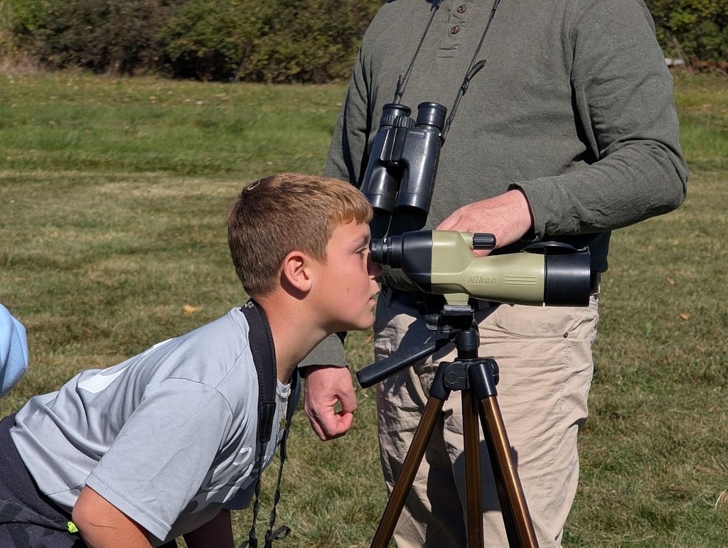 A close-up of a young student leaning forward to look through a tripod-mounted Nikon spotting scope held steady by an instructor.