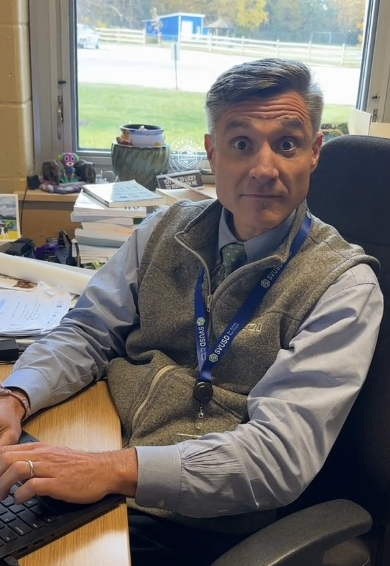 Man sitting at a desk, typing on a keyboard, wide eyed wearing a vest and lanyard