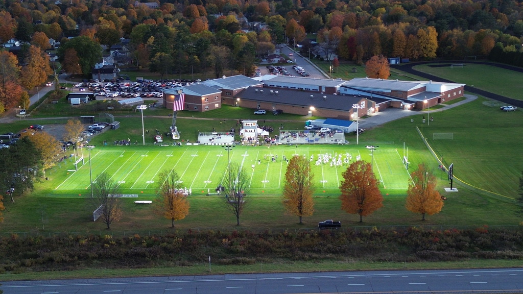 Medium-altitude aerial view of an illuminated football field with players gathered, lined by orange fall trees, bleachers, and a large American flag hanging from a fire truck ladder.