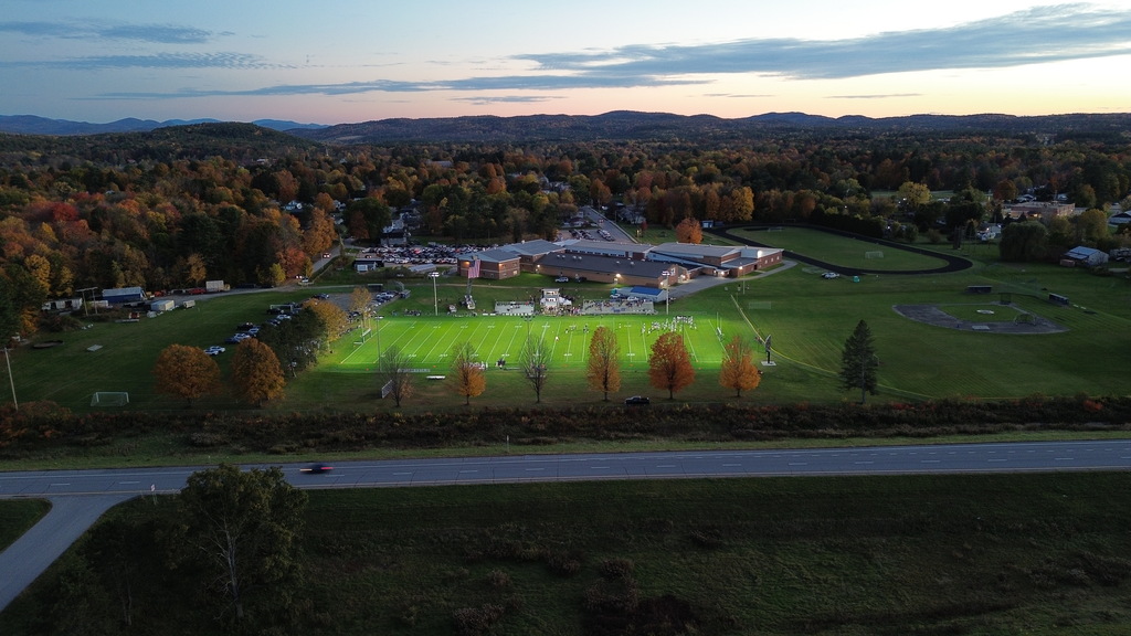 Wide aerial drone shot at dusk showing a brightly lit high school football field surrounded by vivid autumn foliage, high school buildings, and rolling mountains in the distance.