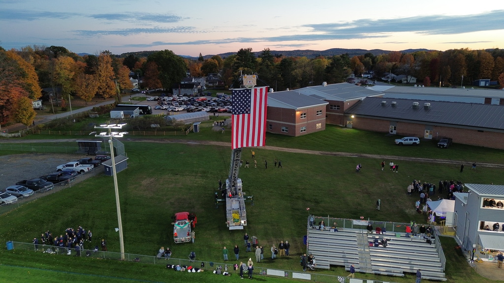 Aerial view focused on a giant American flag suspended vertically from an extended fire department ladder truck next to stadium bleachers and spectators gathered on the sideline.