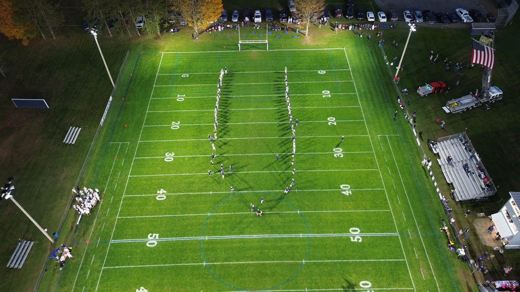 Top-down overhead aerial view directly above a brightly lit green football field, showing two teams of players lined up on opposite sides of the field with bleachers and vehicles around the perimeter.
