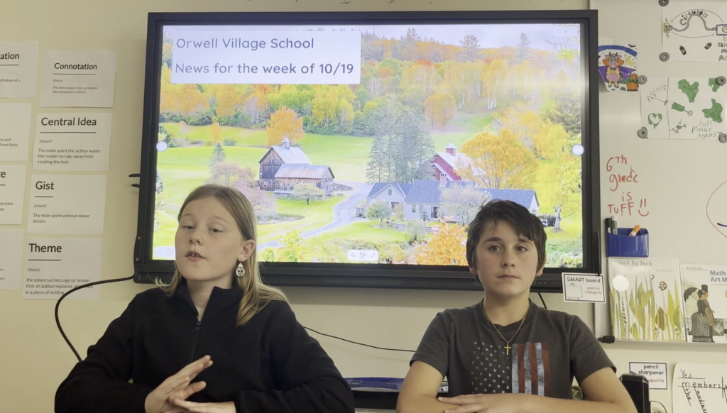 Two Orwell Village School students presenting weekly announcements in a classroom setting. In the background, a large interactive display screen reads "Orwell Village School News for the week of 10/19" set over a scenic autumn photo of a Vermont farm surrounded by fall foliage. To the left, posters define vocabulary terms such as "Connotation," "Central Idea," "Gist," and "Theme".