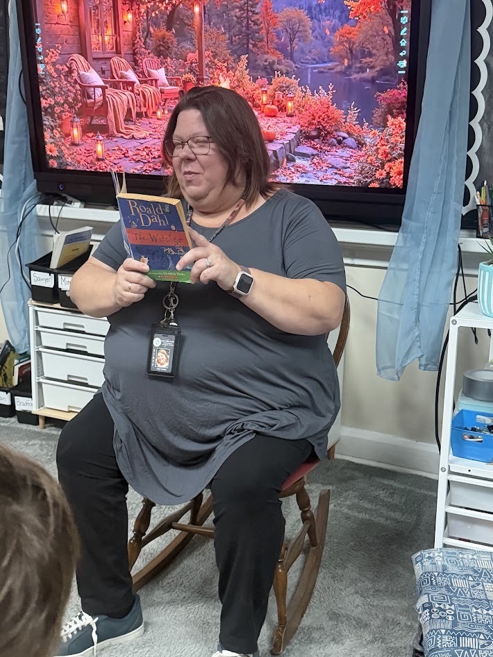 A teacher sitting in a wooden rocking chair in front of a digital display, reading aloud from Roald Dahl's book The Witches.