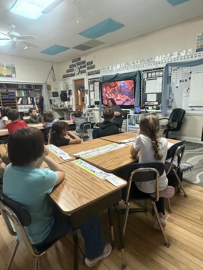 Wide shot of a brightly lit classroom filled with students sitting at desks facing their teacher as she reads aloud from a book.