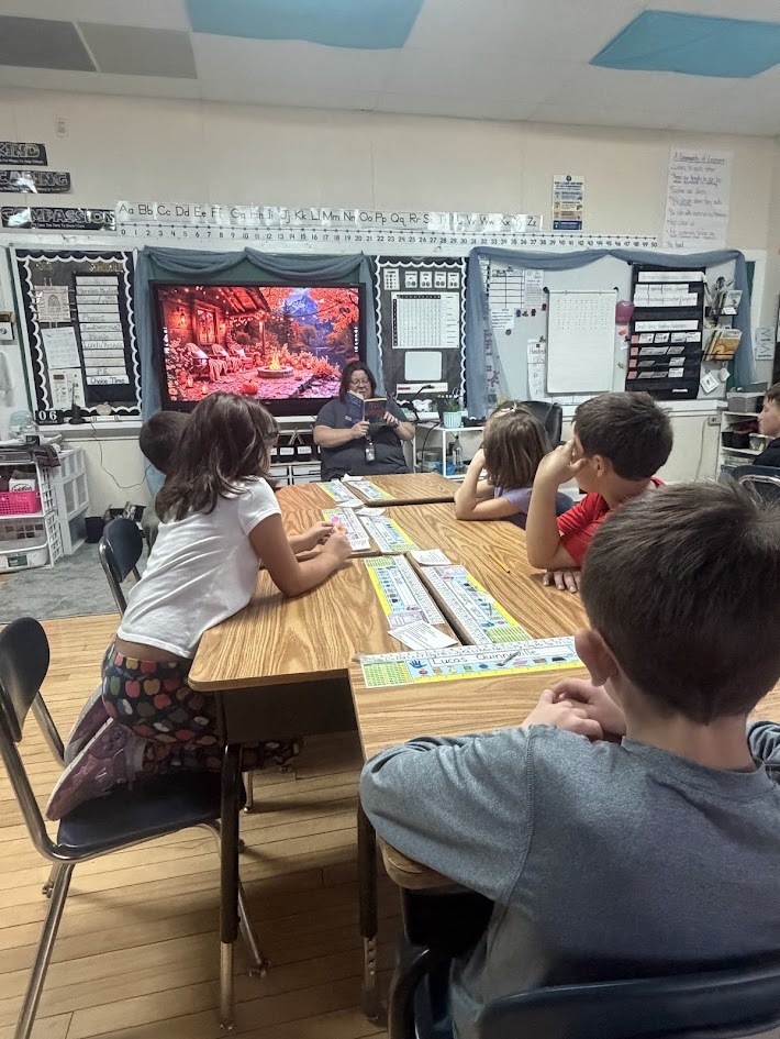 Elementary students seated at wooden desks listening attentively while their teacher reads aloud at the front of the classroom.