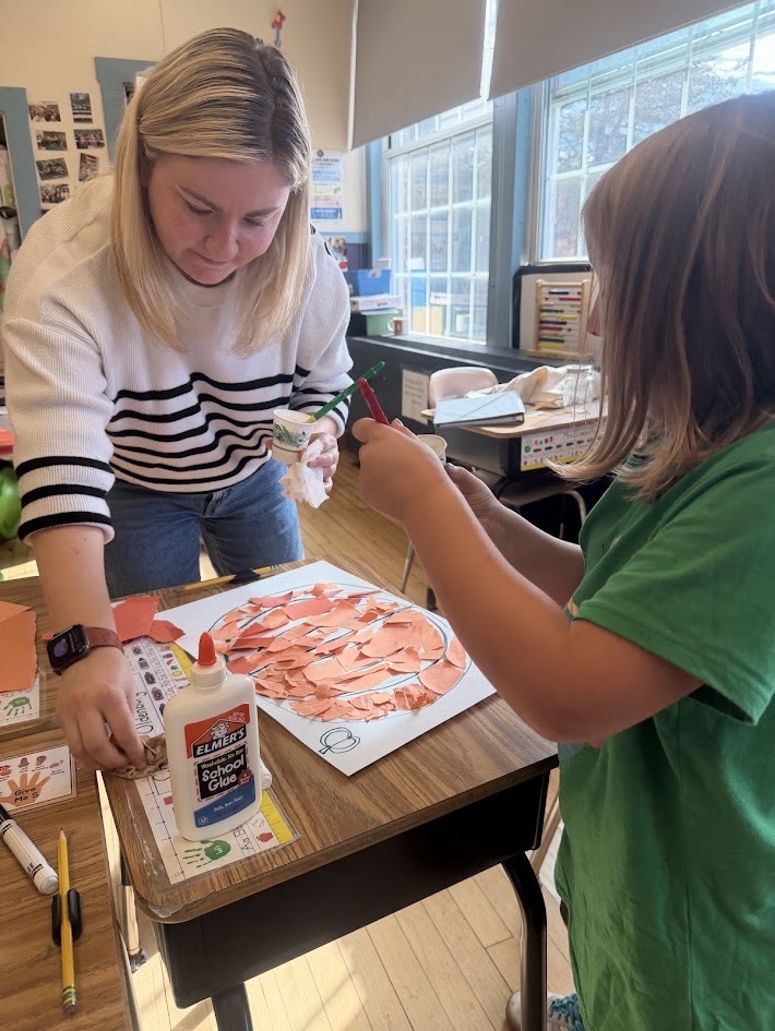A teacher bending down next to a student's desk to assist them with glue and torn paper for their pumpkin craft project.