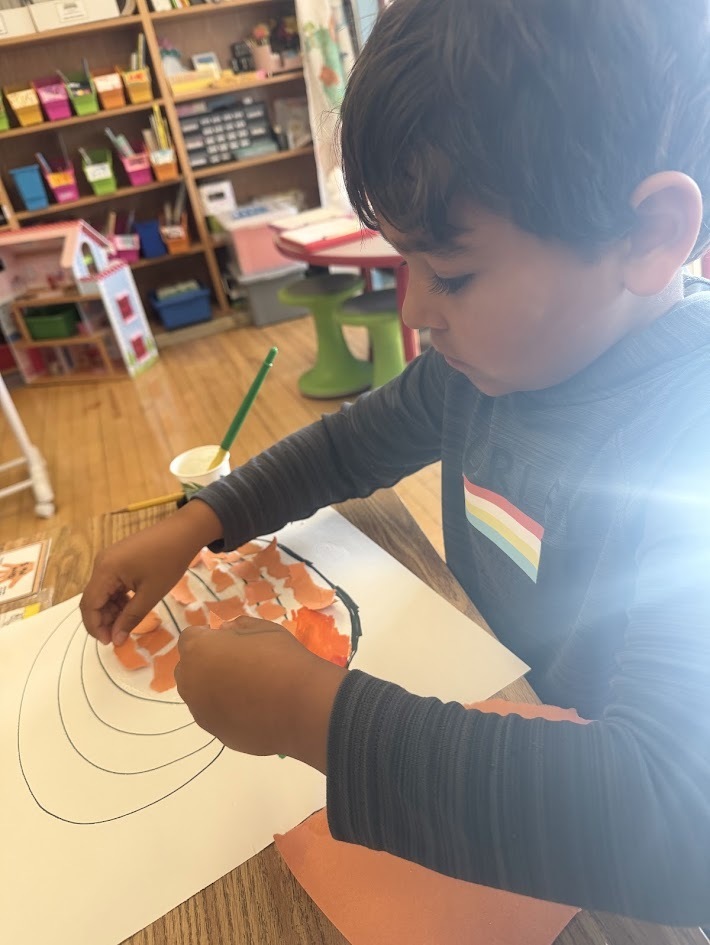Close-up view of a young boy carefully placing orange paper pieces onto a pumpkin outline at his desk.